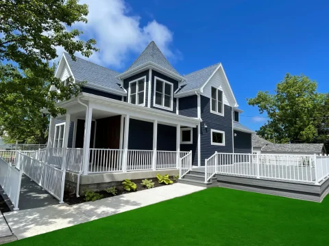 A two-story dark blue house with white trim and a wraparound porch, surrounded by a white railing and green lawn, sits under a blue sky with some clouds and trees nearby.