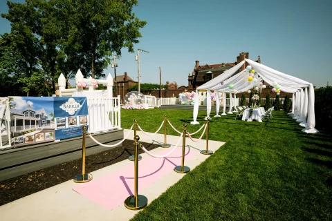 The green space at the Barker Cottage. Velvet ropes lead to a canopy covering a seating table that is topped by flowers.