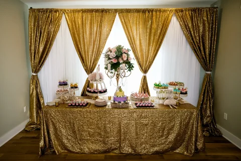 Barker Cottage interior with long fold table against agains a backdrop of gold curtains. The table is covered with flowers, a cake, and cupcakes.