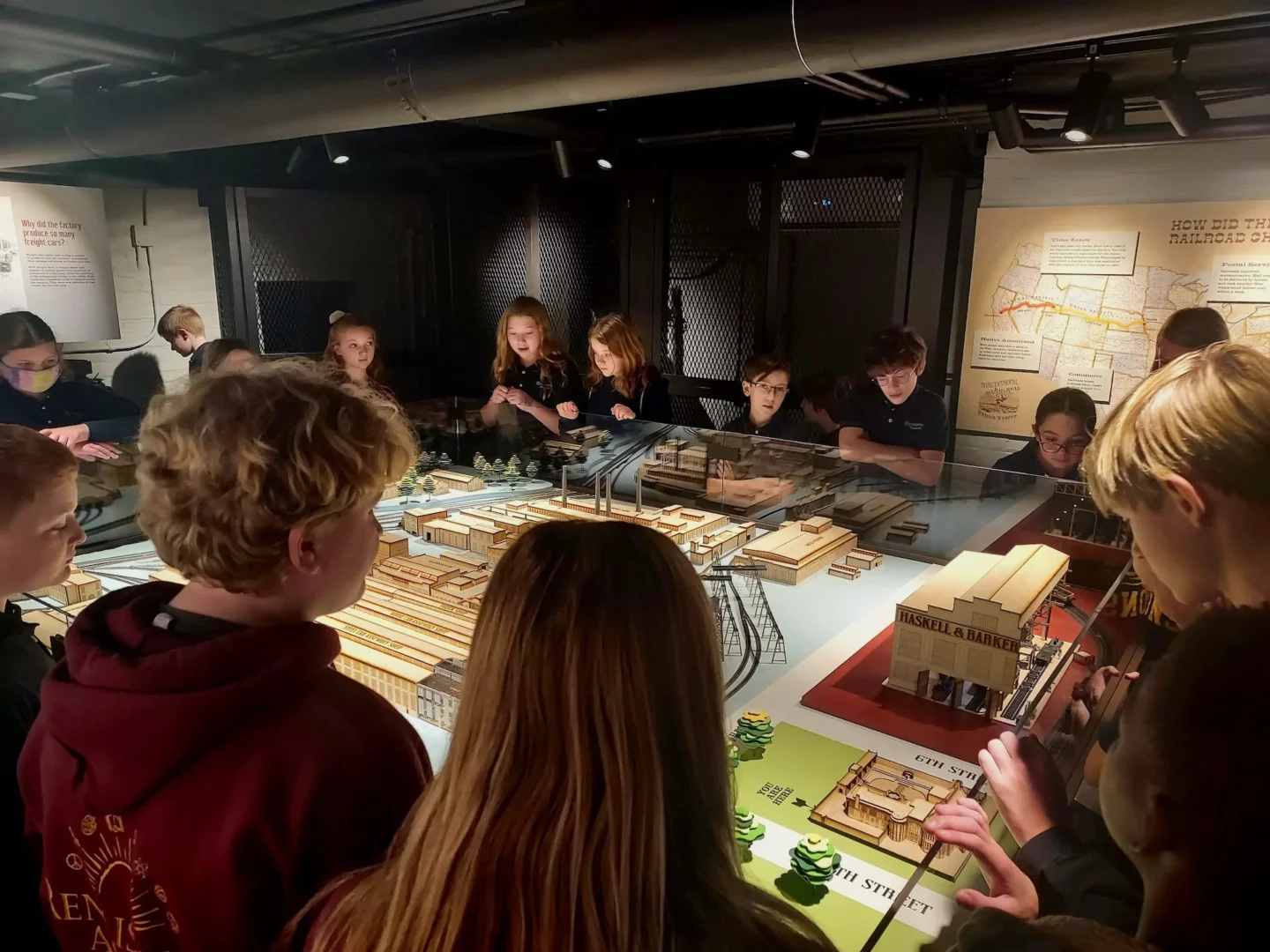 A group of students stand around and observe a detailed architectural model inside a museum exhibit at Barker Mansion in Michigan City, with informational panels and maps displayed on the walls in the background.
