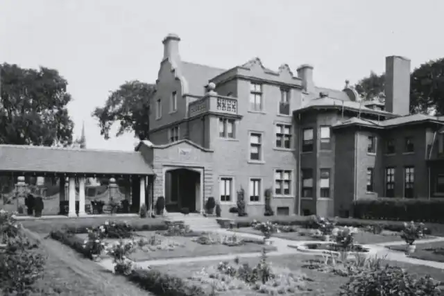 Barker Mansion, a large brick estate in Michigan City, stands behind landscaped gardens, winding pathways, and a covered porch. The garden features vibrant flower beds and neatly trimmed bushes that enhance its historic charm.